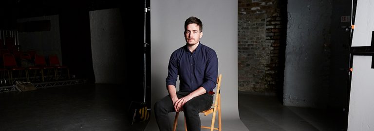 A photo of Tim Foley, a white man, sitting down against a grey wall. He wears an open-necked blue shirt with sleeves rolled up to his elbows, and dark blue trousers. He has short black hair and short facial hair.