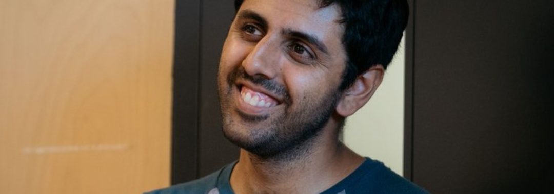 A photo of Vinay Patel, a man of south-Asian heritage. He has short black hair, and smiles broadly, looking off to something in the distance. He wears a teal and grey t-shirt.
