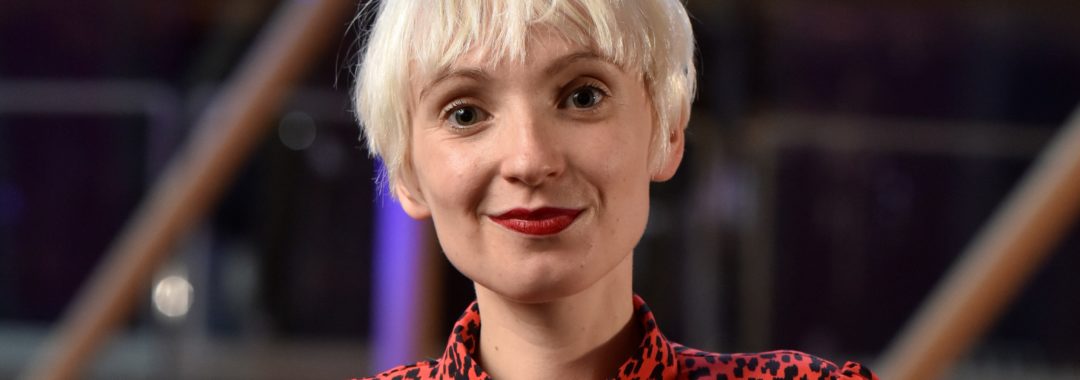A headshot photo of Phoebe Eclair-Powell, a white woman with bleached blonde short hair, wearing a red leopard print zip-up top.